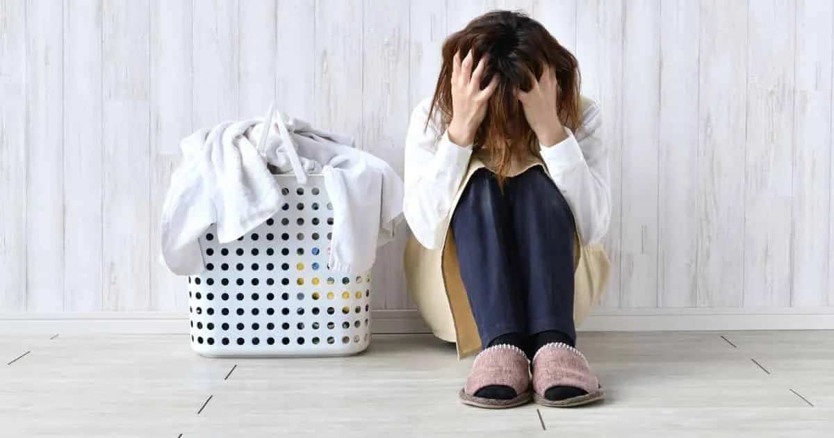 Mère assise à côté d’une panière à linge, visiblement épuisée, illustrant la sensation d’être débordée à la maison et la surcharge mentale domestique