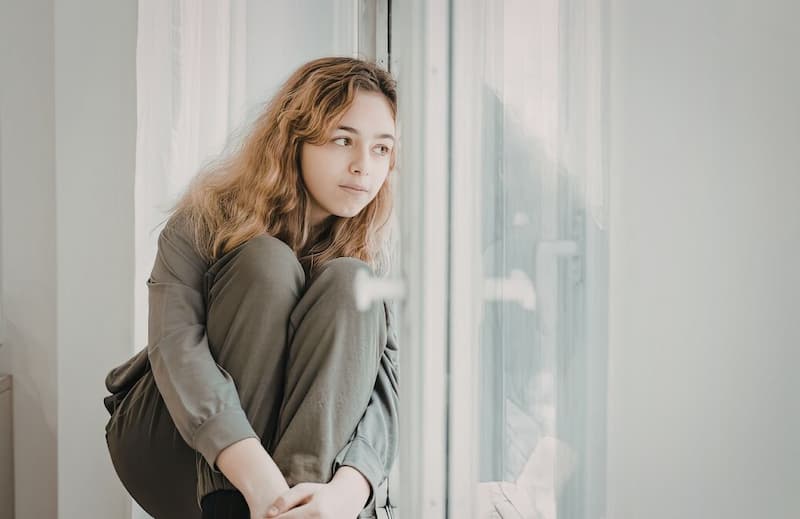 Jeune femme pensive regardant par la fenêtre, illustrant une réflexion sur à quoi sert un sophrologue face au stress et à la charge mentale.